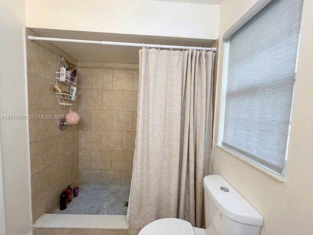 Walk-in shower featuring neutral tile surround, stone-finish flooring, and a recessed threshold, accompanied by a window with a fitted roller shade and a standard white toilet
