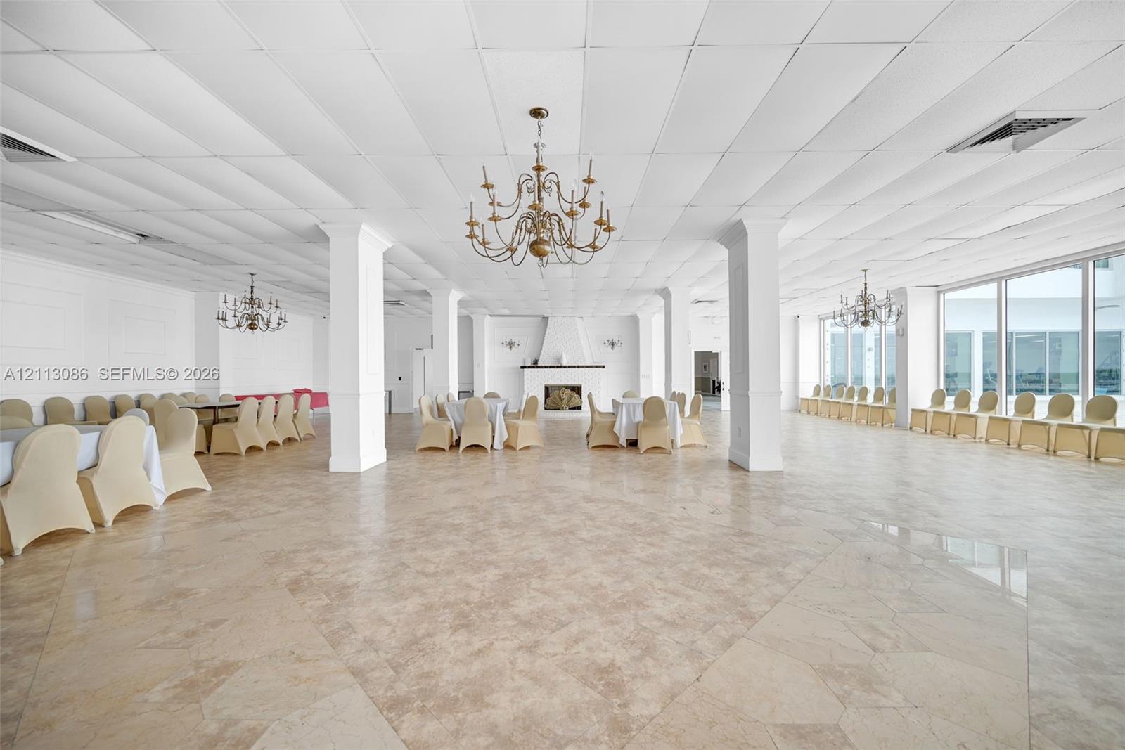Expansive open-concept hall featuring large-format tile flooring, a central fireplace with a decorative mantel, structural columns, ornate multi-arm chandeliers, and floor-to-ceiling windows