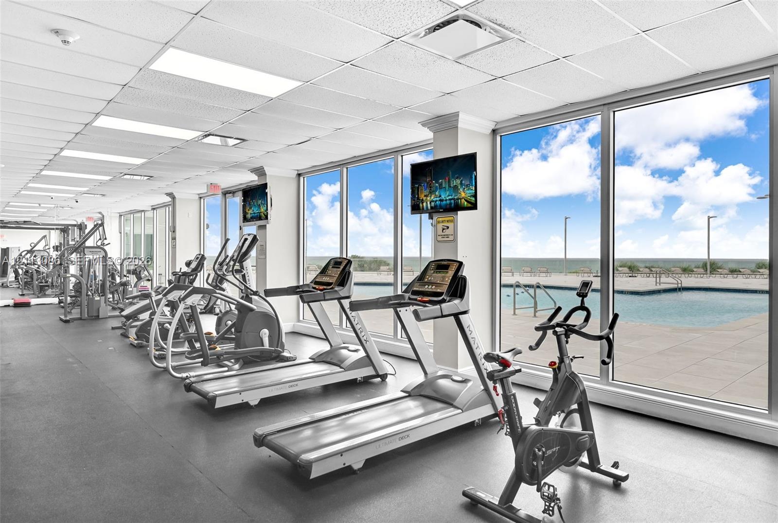 Fitness center featuring wall-to-wall windows, expansive natural light, rubber flooring, drop ceiling panels, and direct sightlines to an outdoor pool