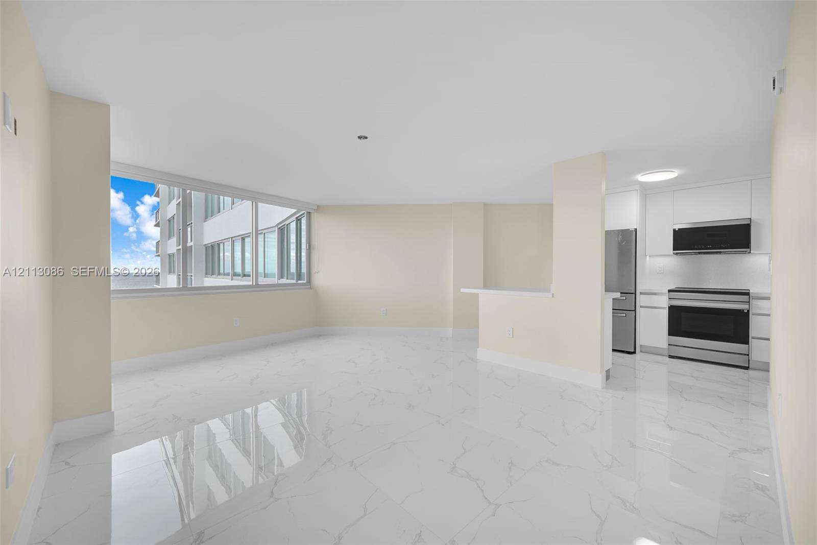 Open-concept living space featuring large-format tile flooring, expansive floor-to-ceiling windows, and a peninsula kitchen with white cabinetry and stainless steel appliances Open-concept living space featuring large-format tile flooring, expansive floor-to-ceiling windows, and a peninsula kitchen with white cabinetry and stainless steel appliances