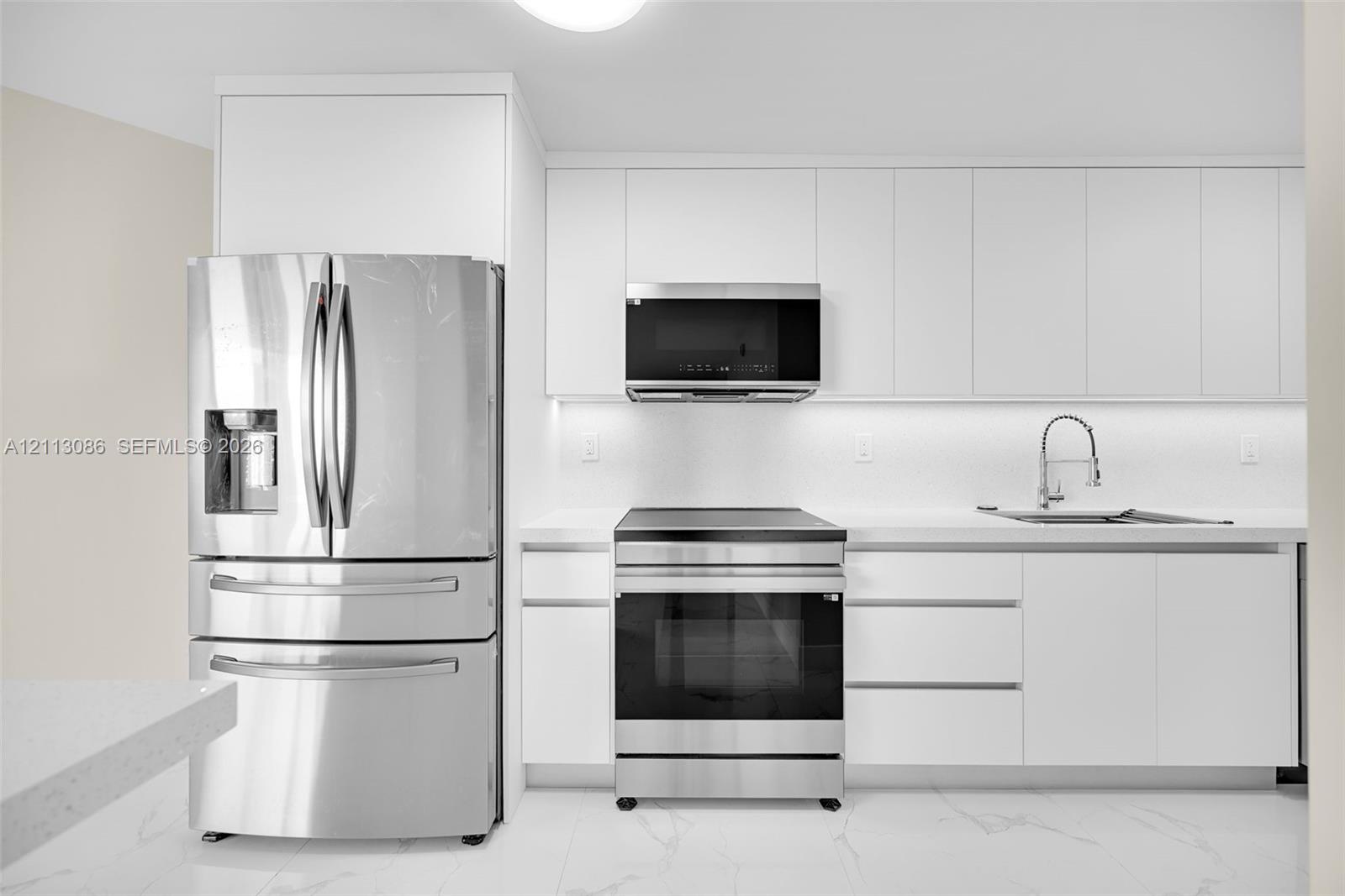 Minimalist kitchen featuring matte white flat-panel cabinetry, stainless steel appliances, a high-arc pull-down faucet, and light stone-patterned flooring