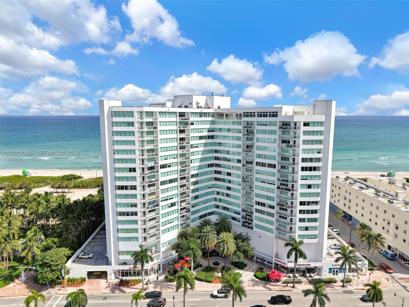 High-rise building featuring a white facade, multiple stacked balconies, floor-to-ceiling glass windows, ground-level covered parking, and an established landscaped courtyard