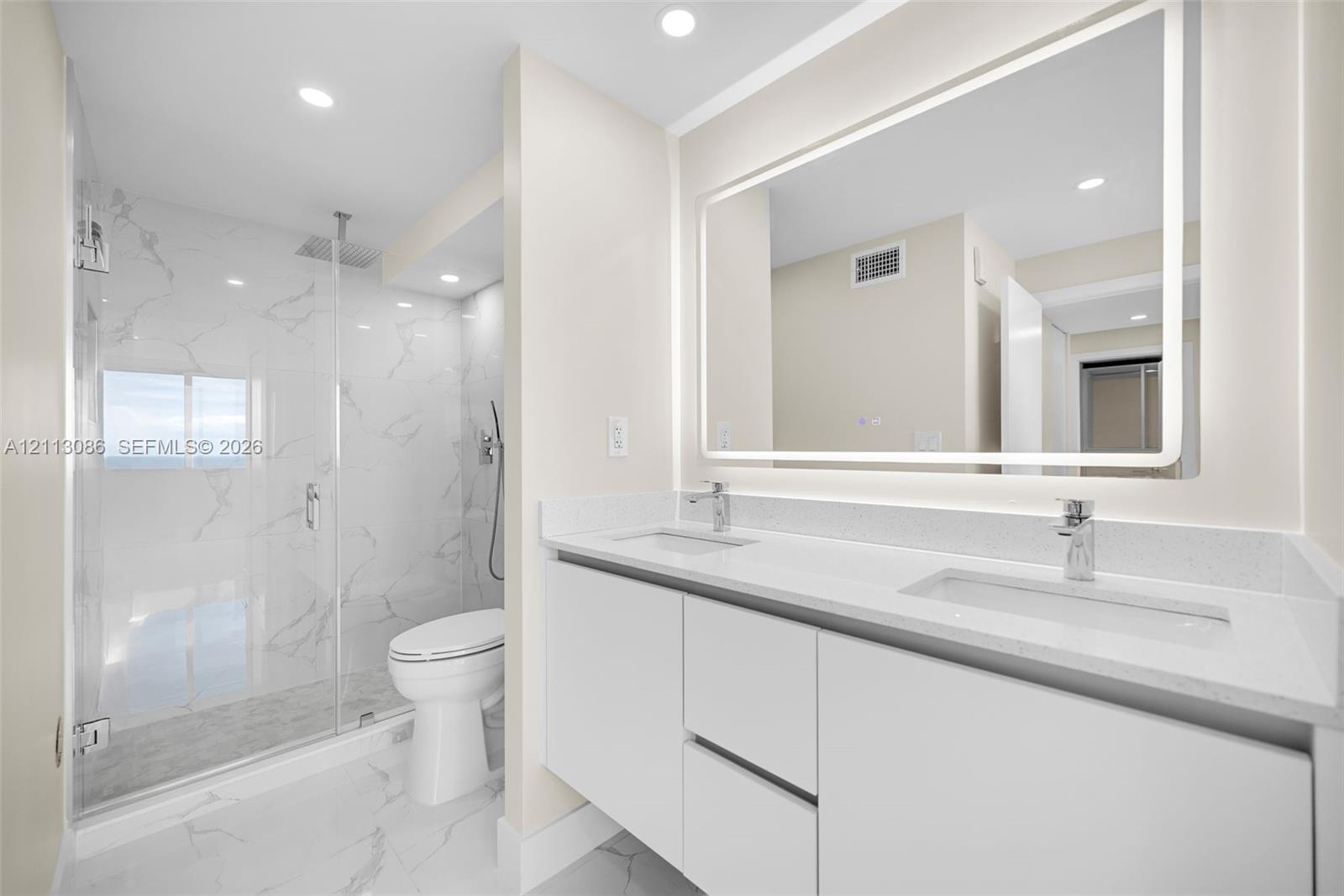 Dual-sink vanity with white cabinetry and solid surface countertop, backlit vanity mirror, walk-in glass shower enclosure featuring rainfall shower head and floor-to-ceiling stone-finish tile surround, and coordinating stone-finish flooring