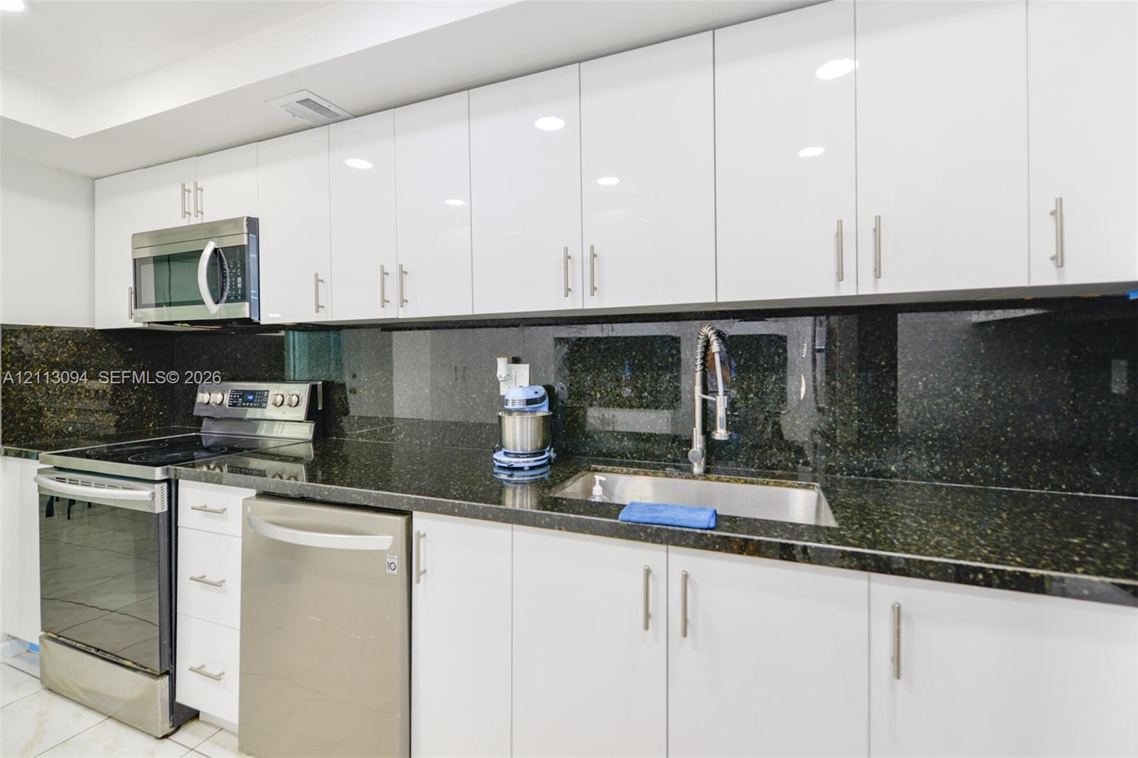 Kitchen featuring high-gloss white cabinetry, dark stone countertops, full-height stone backsplash, stainless steel appliances, and a pull-down faucet