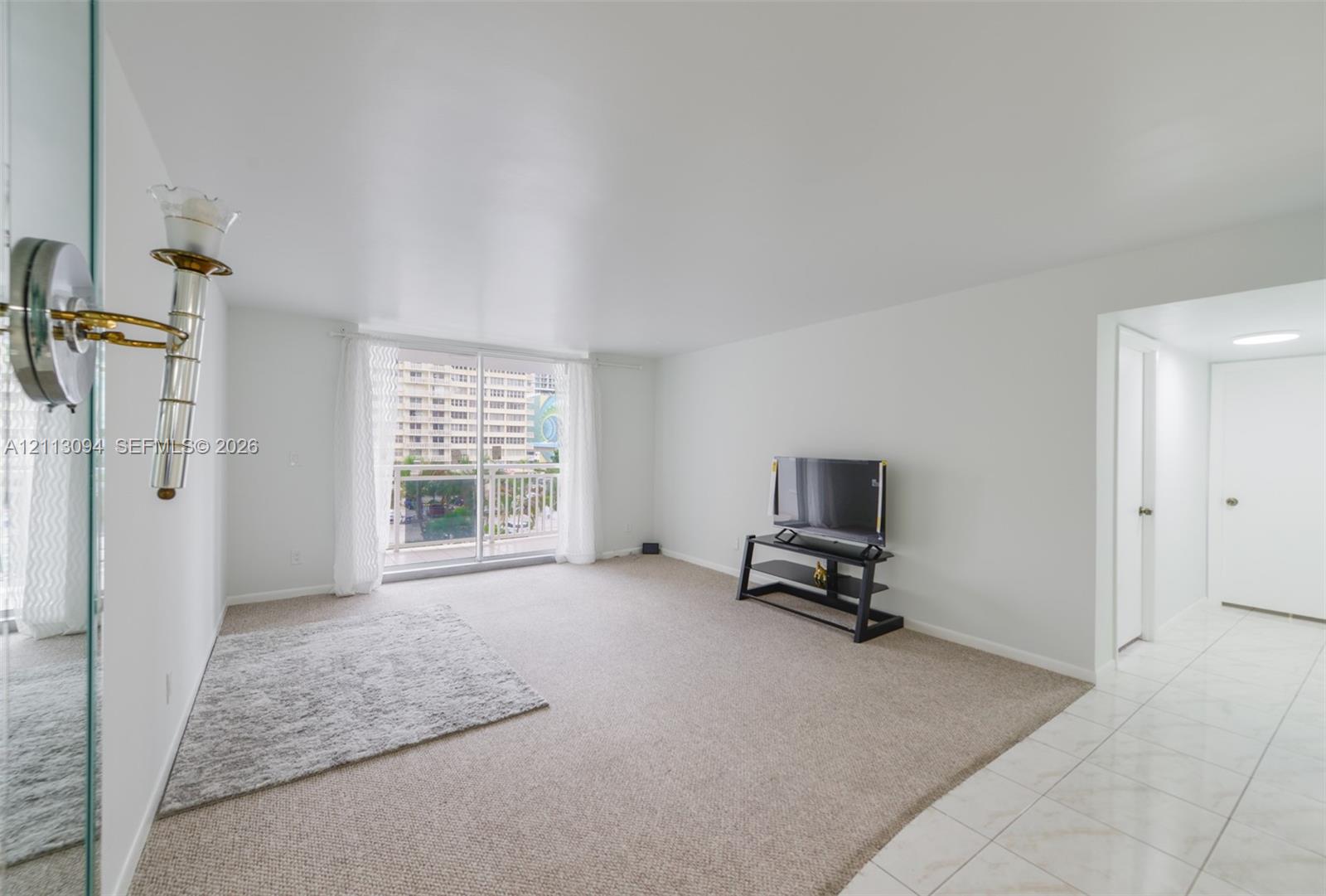 Living area featuring neutral carpeting, ceramic tile flooring, sliding glass door to a balcony, and wall-mounted brass and glass light fixture
