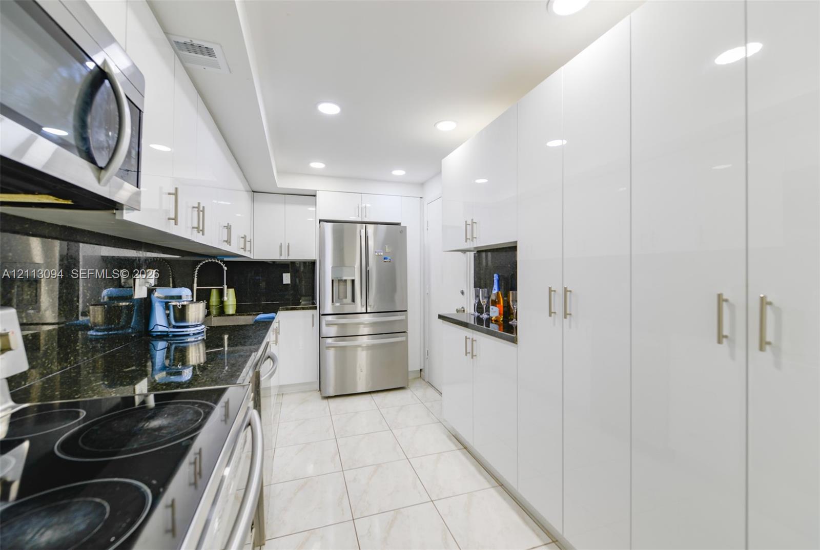 Galley kitchen featuring high-gloss white cabinetry, stainless steel appliances, dark stone countertops, tile flooring, and recessed ceiling lighting