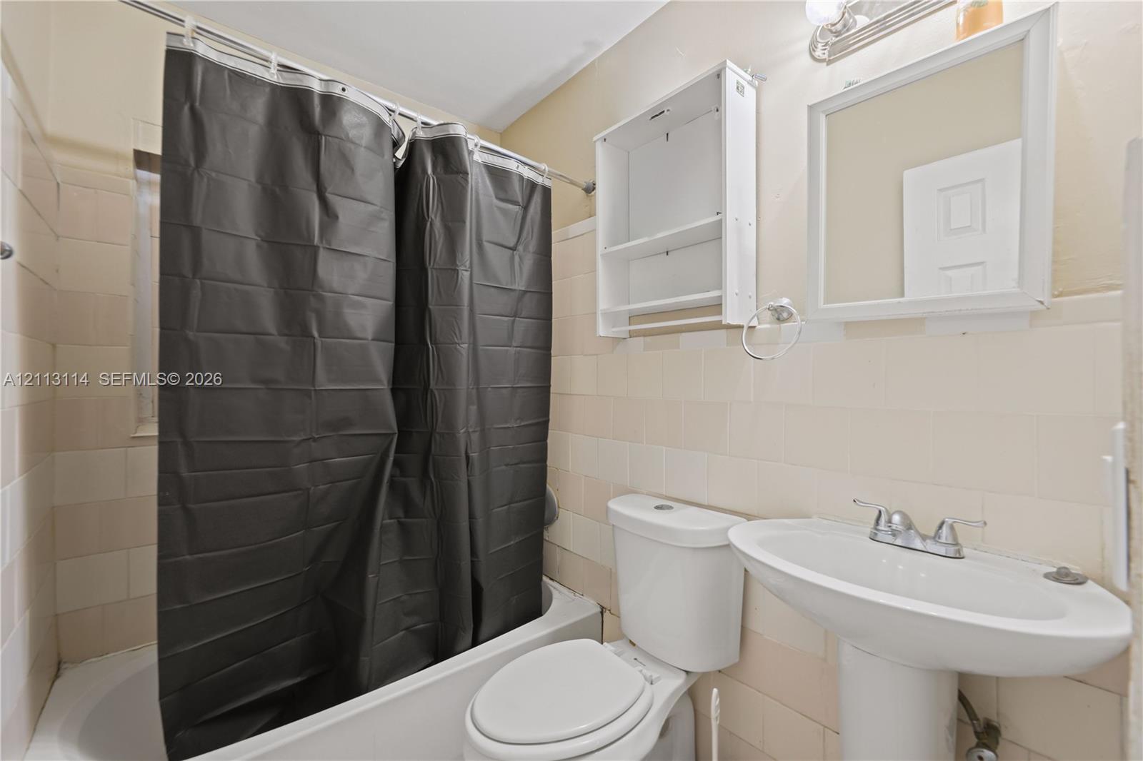Full bathroom featuring a white pedestal sink, a bathtub with wall-mounted plumbing fixtures, neutral ceramic wall tile, a wall-mounted storage cabinet, and a framed vanity mirror
