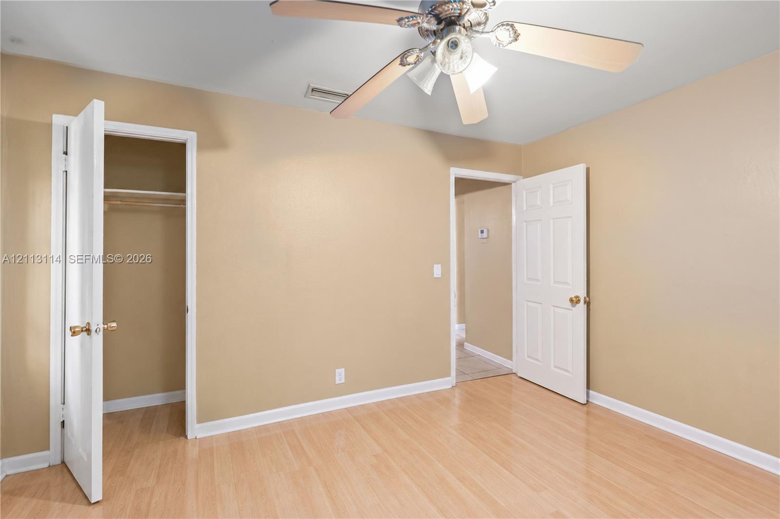 Neutral-toned interior room featuring wood-finish flooring, a multi-blade ceiling fan with integrated lighting, and a reach-in closet with built-in shelving