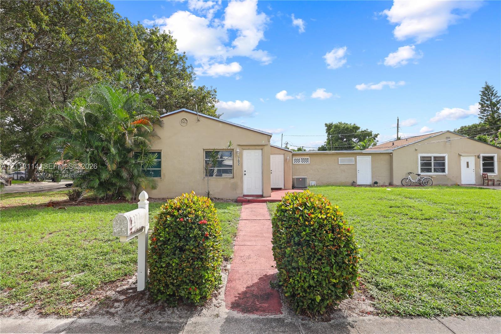 Stucco exterior featuring a straight concrete walkway, manicured shrubbery, mature trees, and a dedicated mailbox