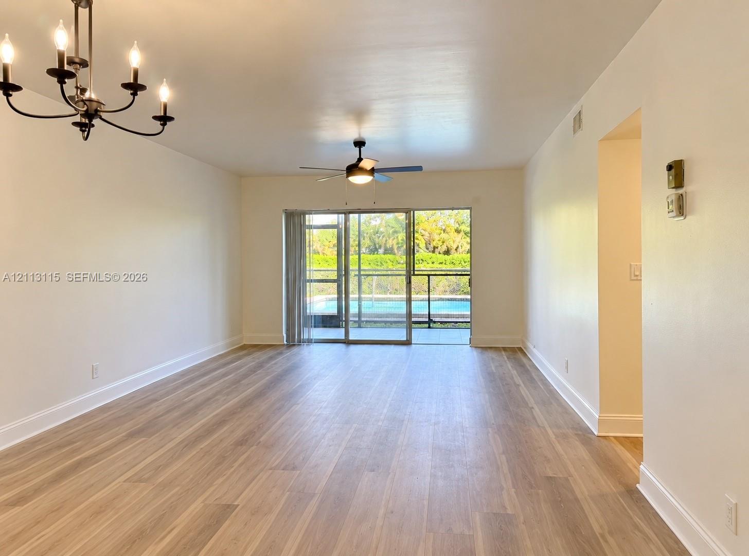 Open-concept interior featuring light wood-finish flooring, a multi-arm iron chandelier, a ceiling fan, and large sliding glass doors providing outdoor access Open-concept interior featuring light wood-finish flooring, a multi-arm iron chandelier, a ceiling fan, and large sliding glass doors providing outdoor access