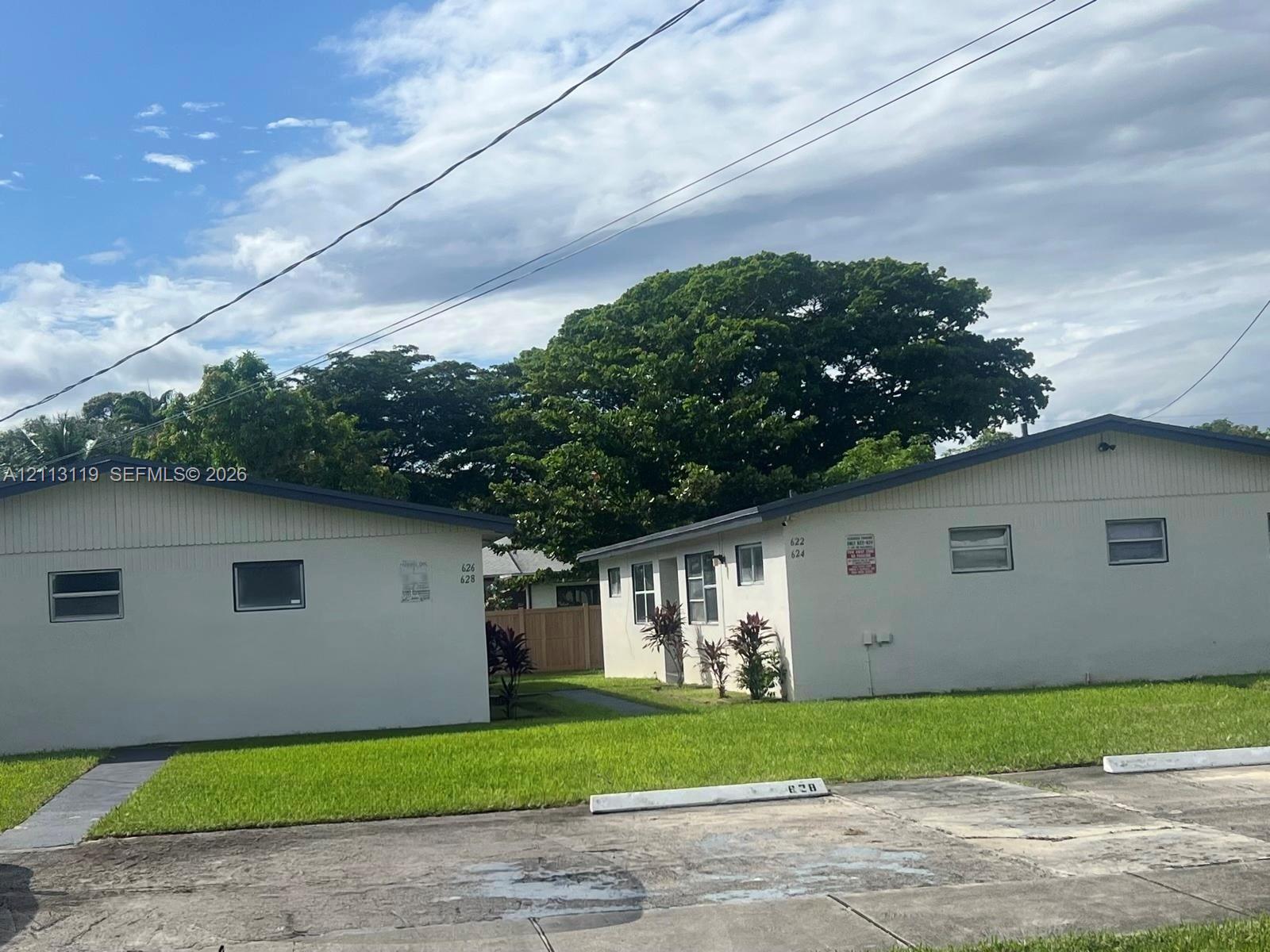 Single-story multi-unit structure featuring a light-toned stucco exterior, asphalt shingle roofing, paved surface parking with concrete bumpers, and a lawn