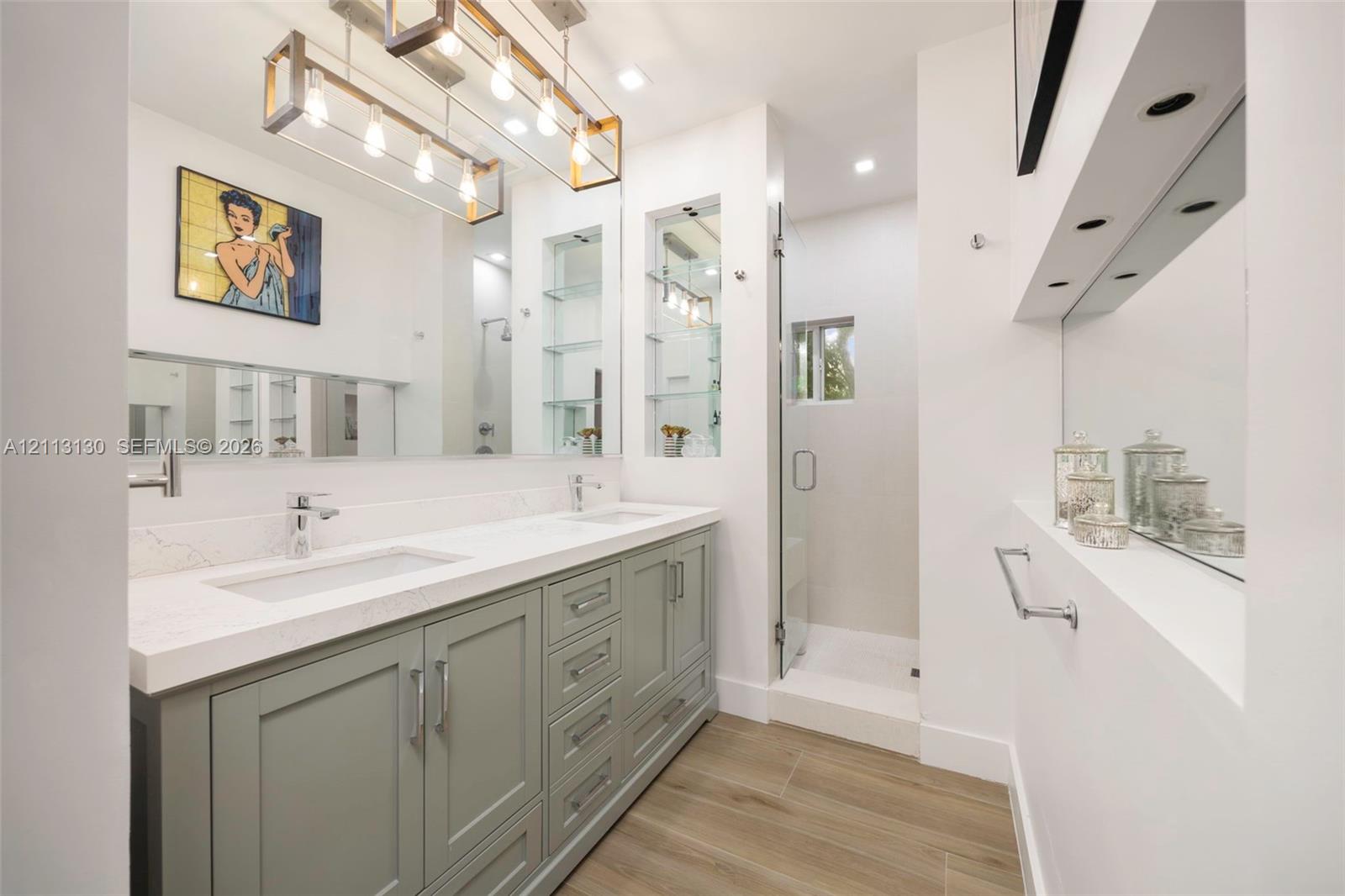 Dual-sink vanity with light gray shaker-style cabinetry, white solid-surface countertop, rectangular undermount sinks, and polished chrome fixtures, complemented by recessed lighting, a linear metal chandelier, and wood-finish flooring