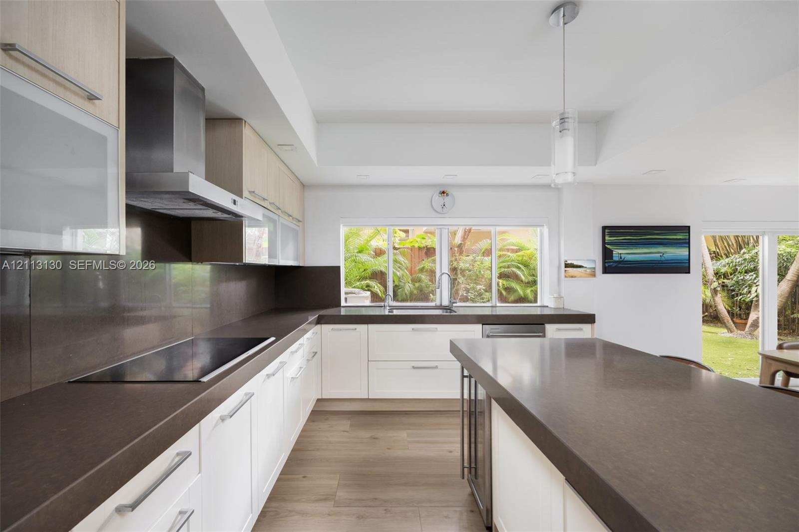 Modern kitchen featuring white flat-panel cabinetry, dark stone-finish countertops, stainless steel range hood, built-in cooktop, and wood-finish flooring