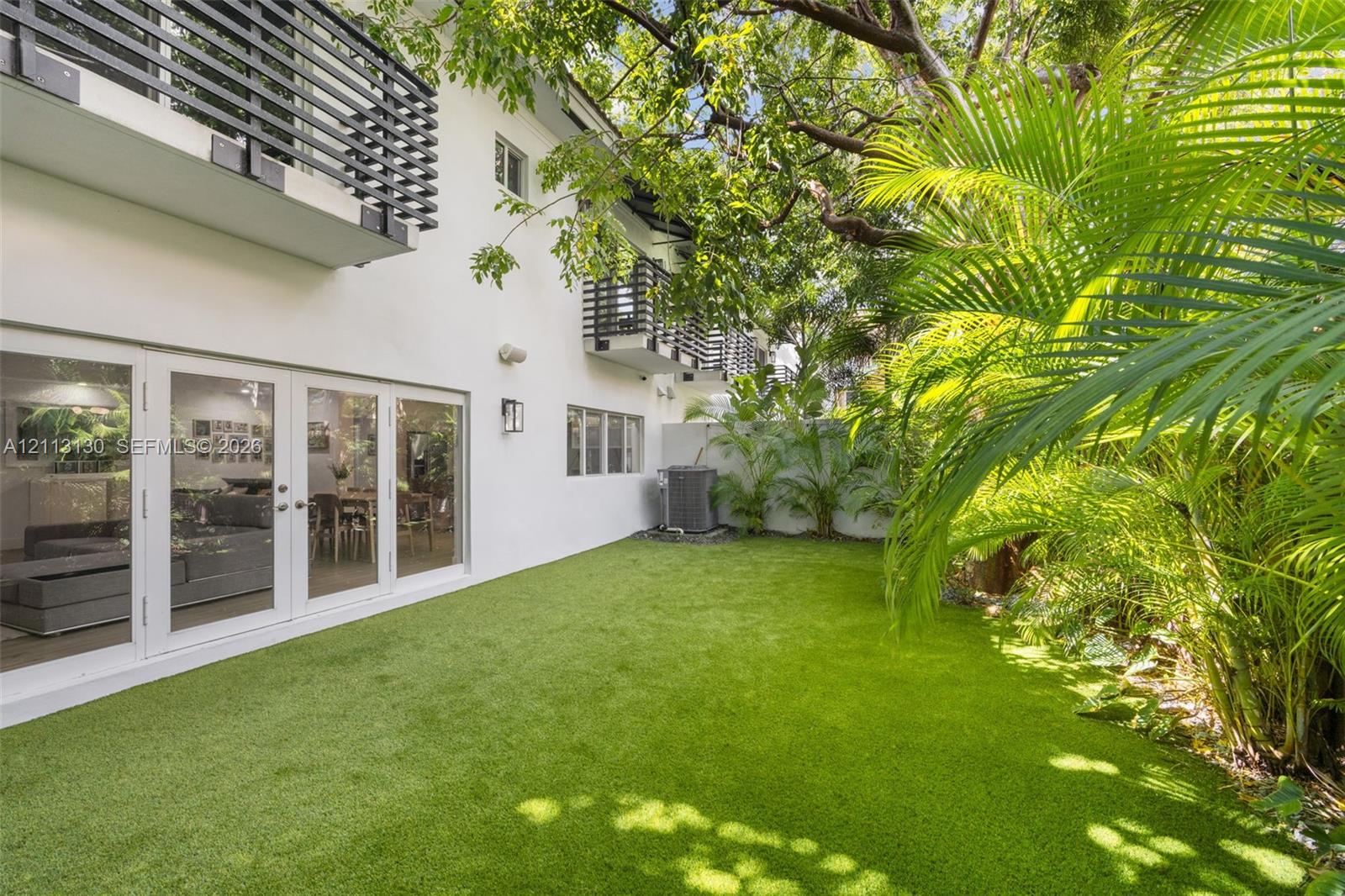 Backyard featuring artificial turf, white stucco exterior, multiple second-story balconies with horizontal railings, and sliding glass panel doors