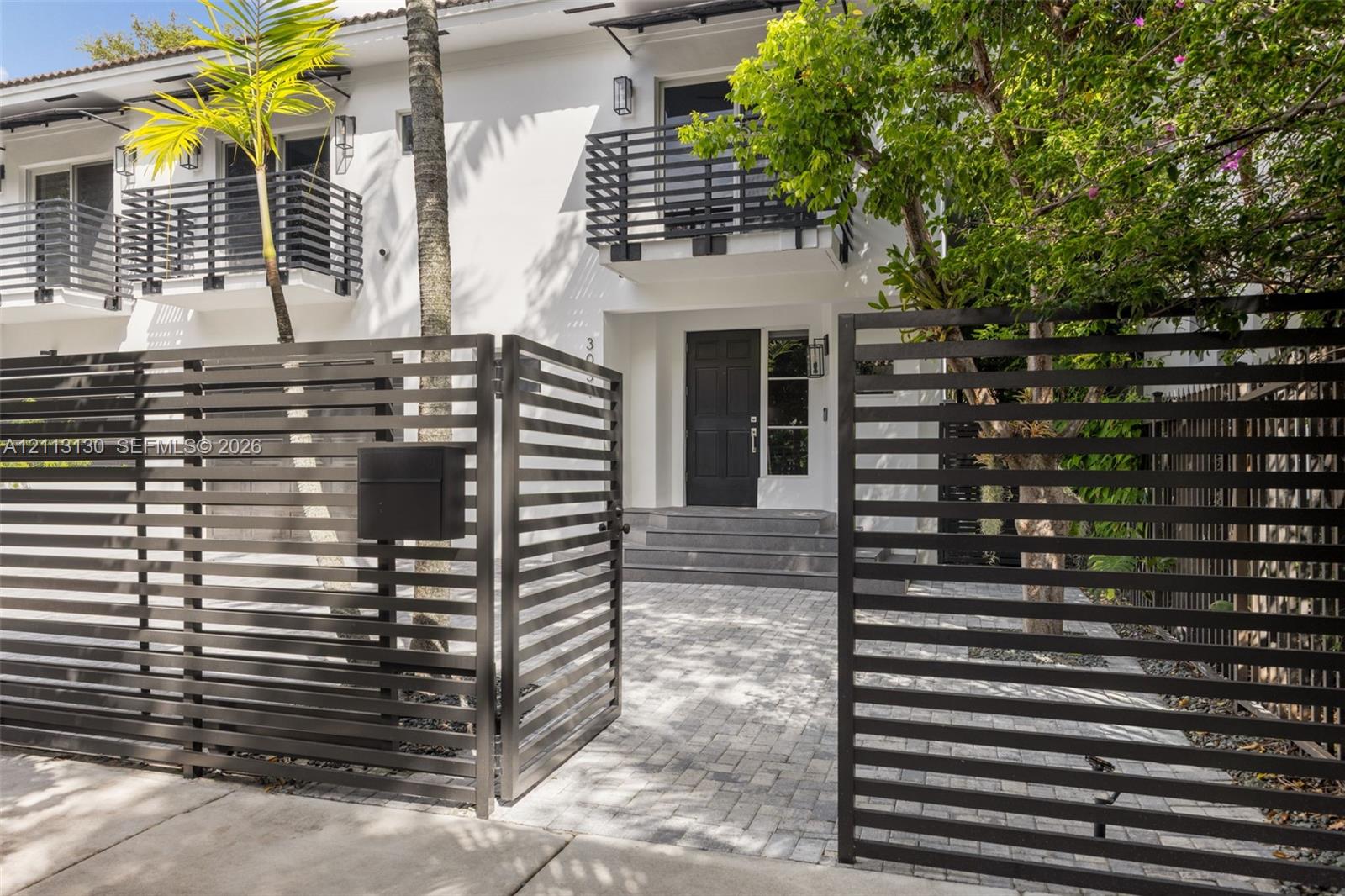 Exterior facade featuring a paved stone driveway, horizontal slat metal privacy fencing, solid panel entry door, and upper-level balconies with matching metal railings