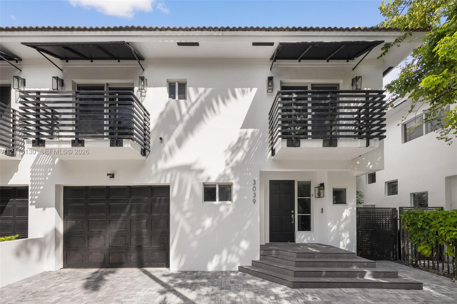 Two-story stucco exterior featuring horizontal black metal balcony railings, a multi-paneled garage door, geometric entry steps, and matching black metal awnings Two-story stucco exterior featuring horizontal black metal balcony railings, a multi-paneled garage door, geometric entry steps, and matching black metal awnings