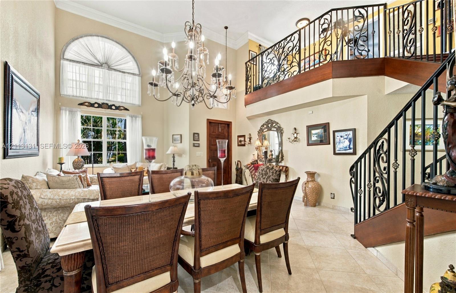 High-ceilinged room featuring an arched window, a prominent chandelier, tile flooring, and an elevated balcony with decorative metal railing
