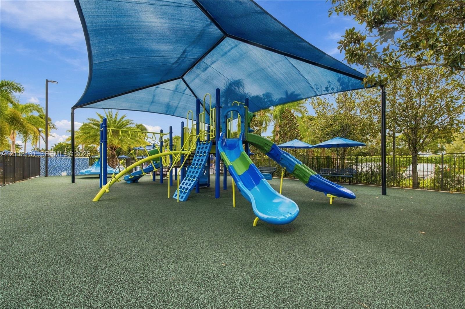 Outdoor recreational area featuring a playground structure with multiple slides, overhead sun shade canopy, and impact-absorbing flooring