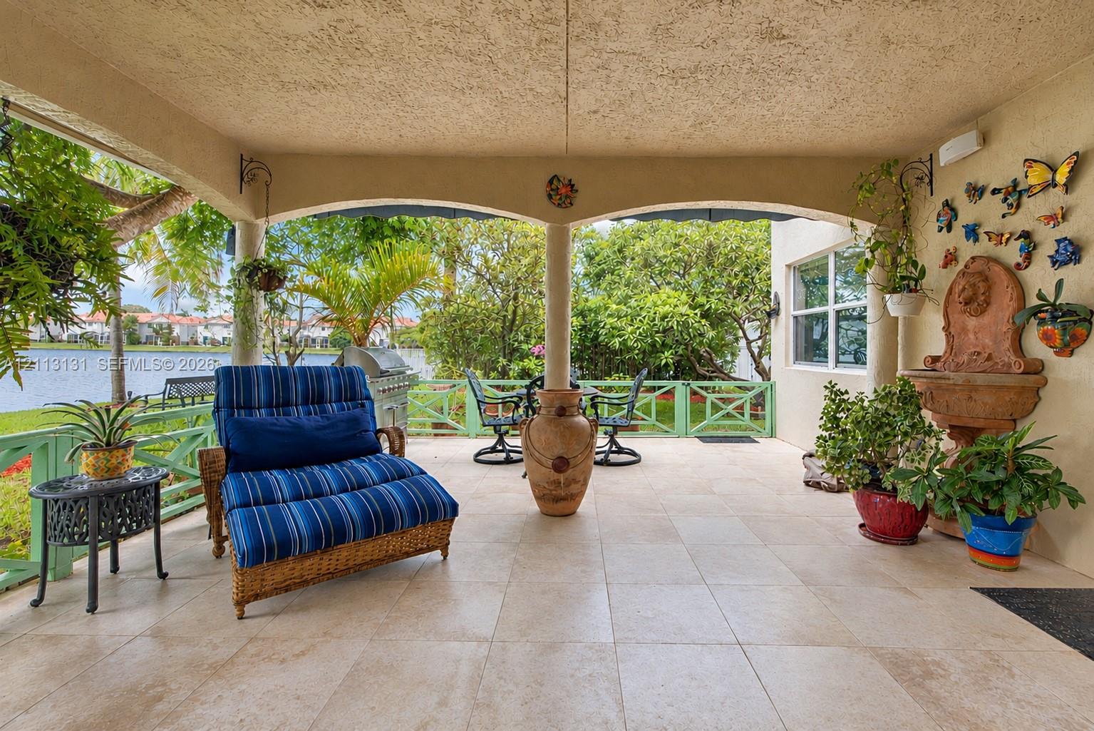 Covered patio featuring stone-tile flooring, arched architectural supports, and a decorative wall-mounted fountain, enclosed by a green lattice railing overlooking the water