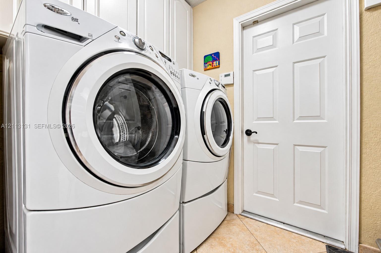 Laundry area featuring built-in white cabinetry, full-size front-load washer and dryer, tile flooring, and a six-panel door with matte black hardware