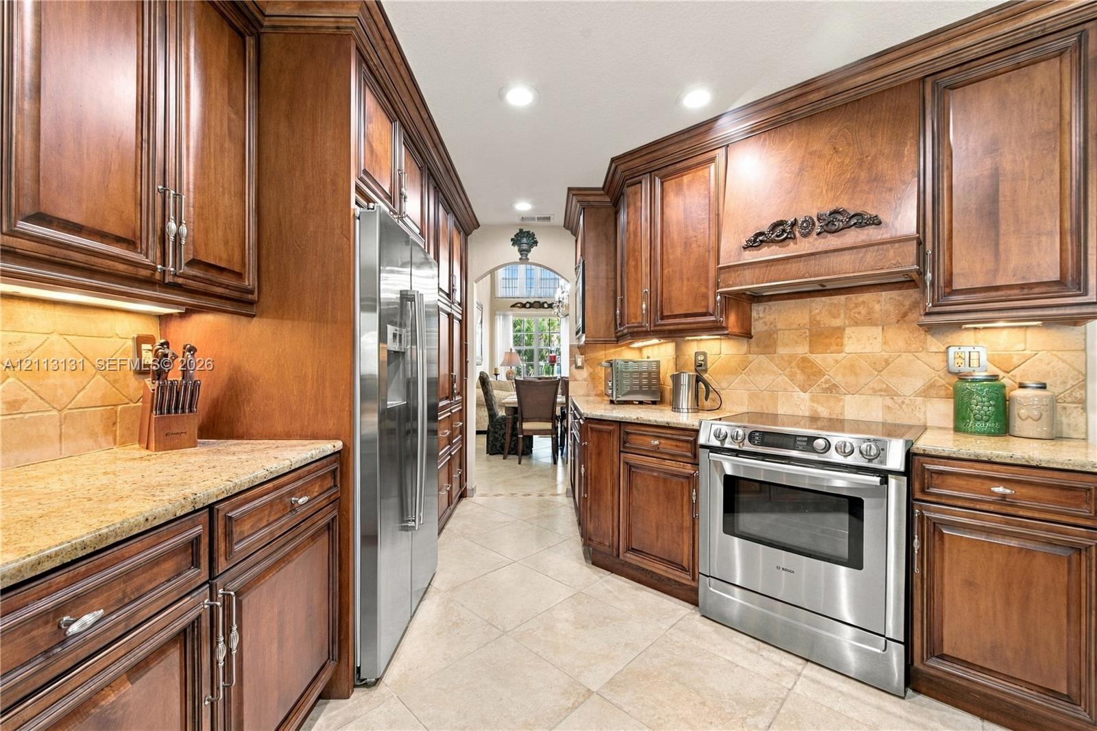 Kitchen featuring custom stained-wood cabinetry, stone-finish countertops, diagonal tile backsplash, stainless steel appliances, and tile flooring