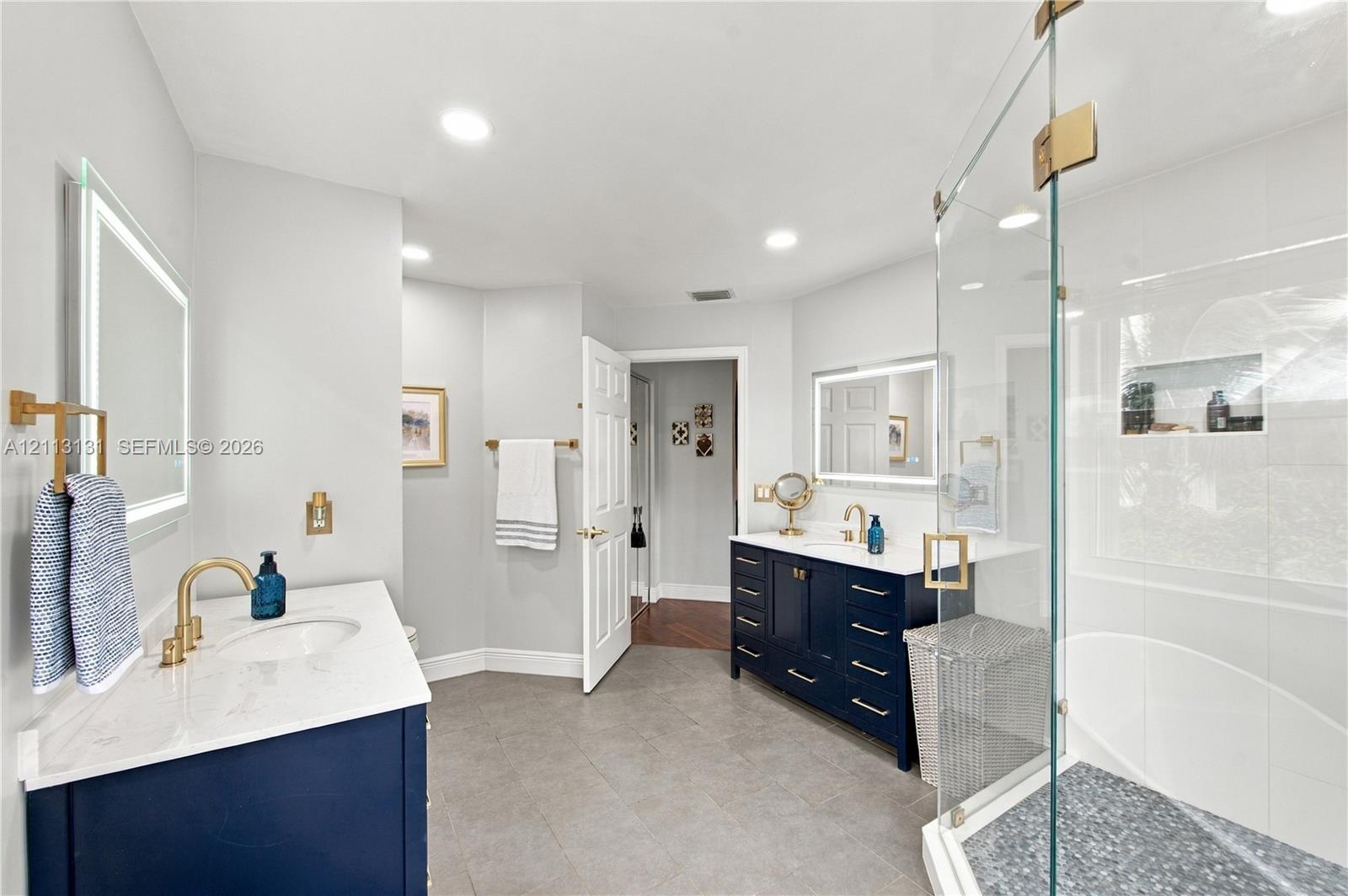 Dual navy blue vanities with white quartz countertops, gold-tone fixtures, illuminated mirrors, and a glass-enclosed shower with a built-in recessed niche and stone-tile flooring