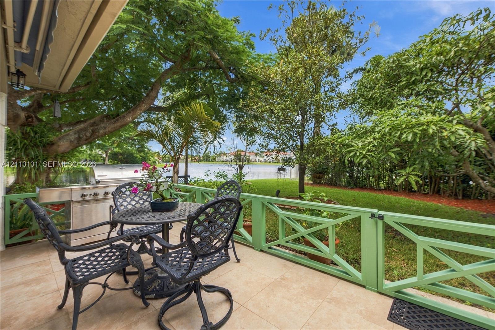 Tiled patio featuring a built-in outdoor grill, painted wood-style railing, and mature trees overlooking a waterfront area