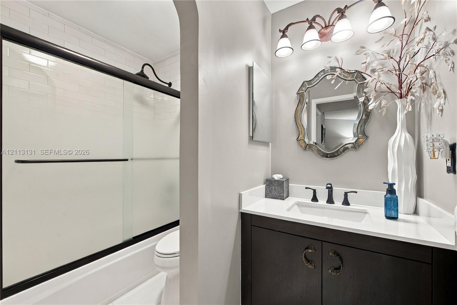 Dark-finish vanity with white countertop, integrated rectangular sink, matte black widespread faucet, and shower featuring a glass enclosure with black framing and white subway tile surround