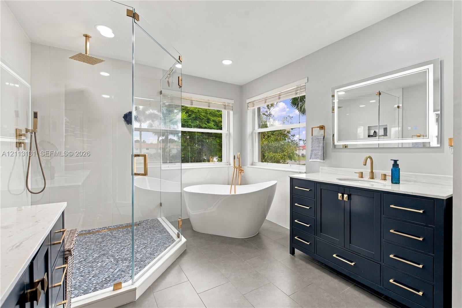 Bathroom featuring a glass-enclosed walk-in shower with pebble-stone flooring, a freestanding soaking tub, a dark-finished dual-drawer vanity with a white stone countertop, and a backlit wall mirror