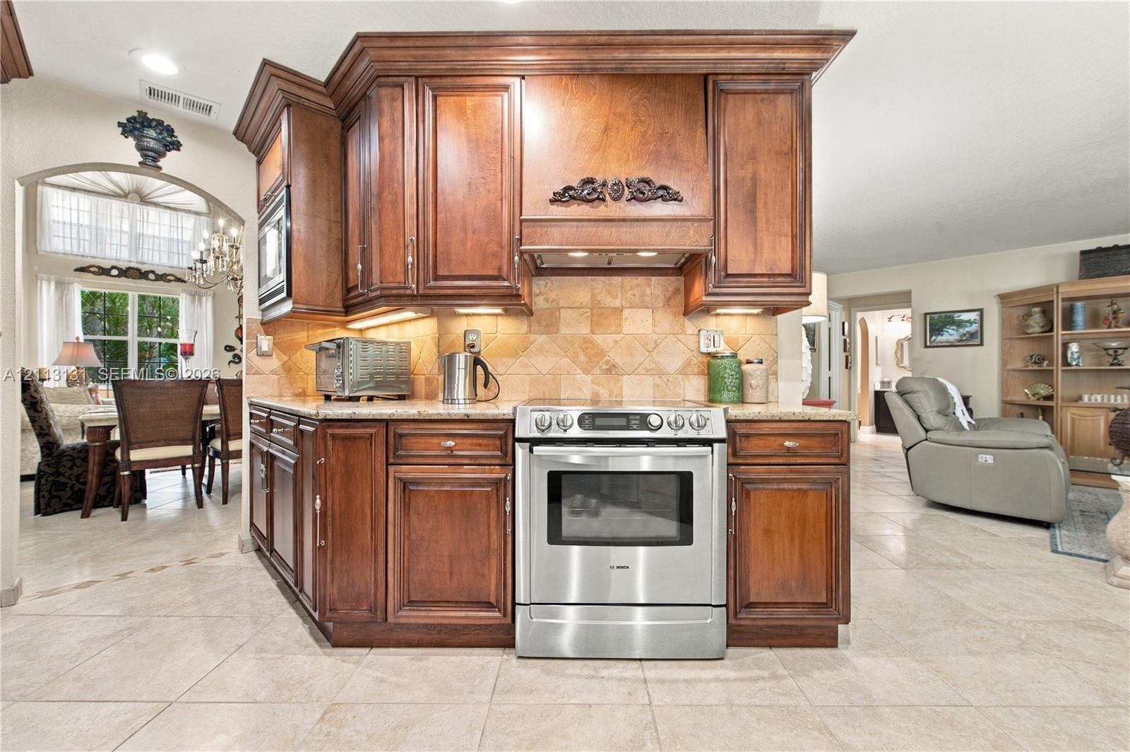 Custom wood cabinetry featuring an integrated range hood, stainless steel oven, stone-tile backsplash, and solid surface countertops with tile flooring