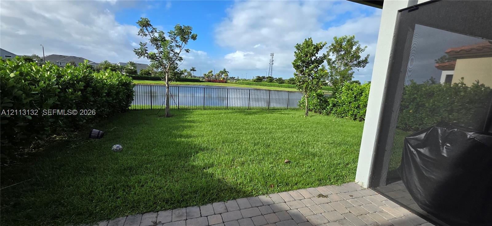 Backyard featuring a paved patio, manicured lawn, perimeter hedge fencing, and metal enclosure overlooking a pond