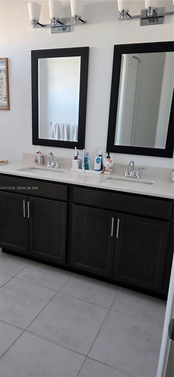Double-sink vanity featuring dark wood cabinetry, solid-surface countertop with integrated basins, chrome hardware, matching framed mirrors, and dual-light wall fixtures over large-format tile flooring