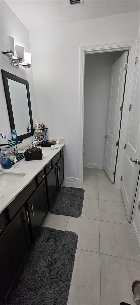 Double-sink vanity featuring dark cabinetry, light stone-finish countertops, three-light wall-mounted fixture, and oversized ceramic tile flooring