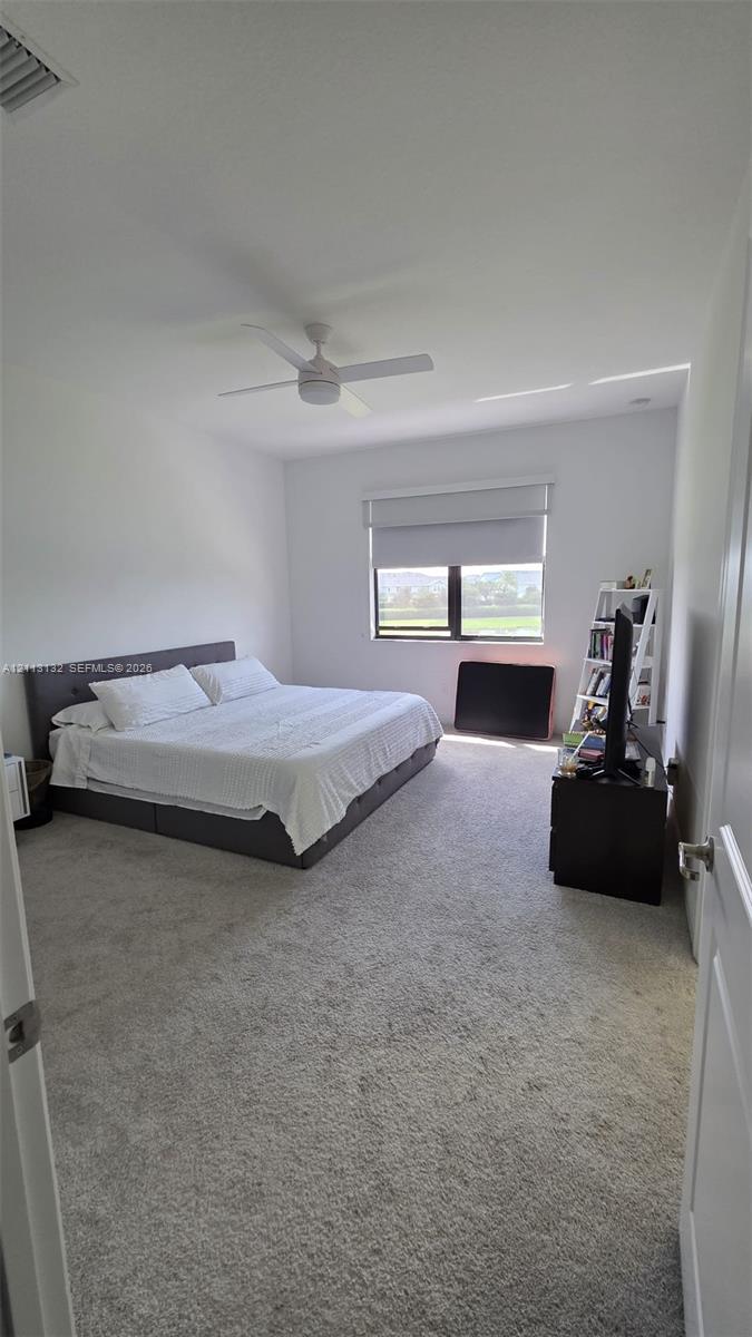 Neutral-toned wall-to-wall carpeting, ceiling fan with integrated lighting, and single-pane window with a roller shade