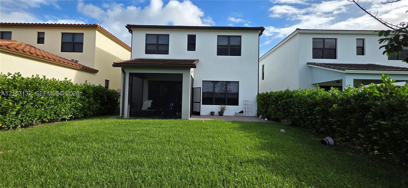 Two-story stucco exterior featuring a screened-in rear patio, concrete hardscaping, and a grass lawn enclosed by mature hedges