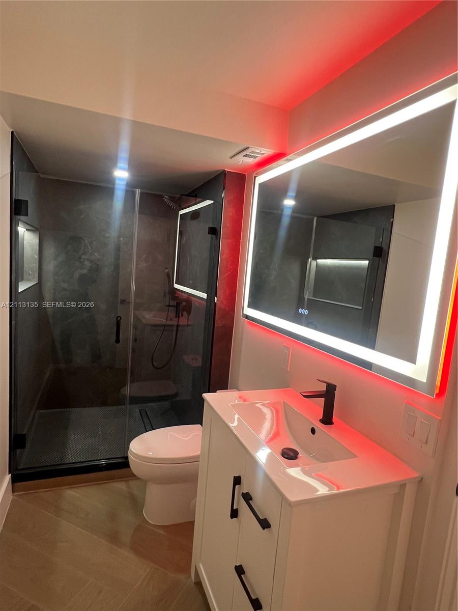 Bathroom featuring a vanity with white cabinetry and matte black hardware, integrated sink with a matte black faucet, wall-mounted LED-backlit mirror, glass-enclosed shower with dark tile surround, and tile flooring