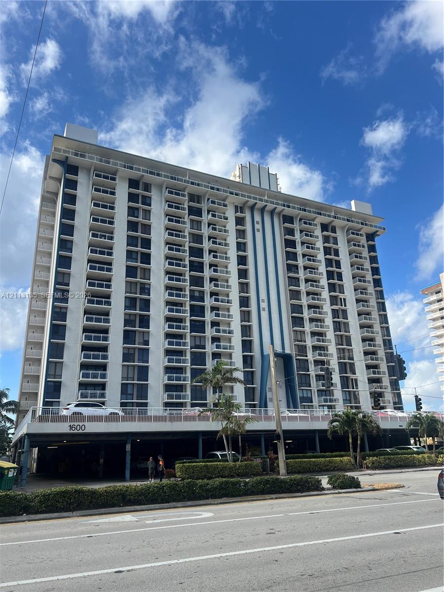 High-rise residential exterior featuring multiple tiers of balconies with metal railings, structural blue-toned architectural accents, and a covered ground-level parking area High-rise residential exterior featuring multiple tiers of balconies with metal railings, structural blue-toned architectural accents, and a covered ground-level parking area