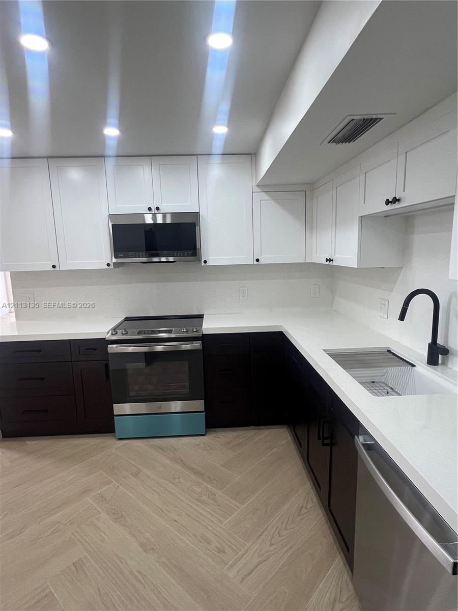 Kitchen featuring two-tone cabinetry, white stone-composite countertops, stainless steel appliances, matte black hardware, and wood-finish herringbone flooring