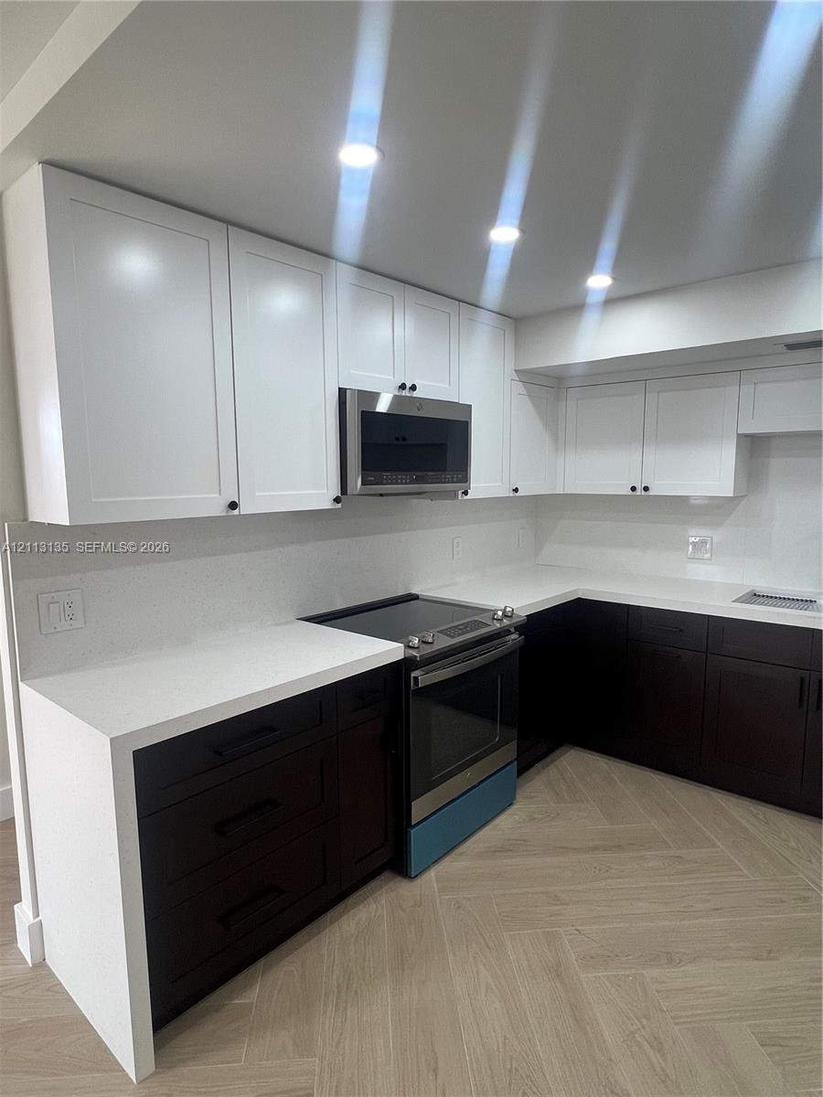Two-tone cabinetry featuring white upper cabinets and dark lower drawers, solid surface countertops, stainless steel over-the-range microwave, electric range, and wood-finish flooring in a herringbone pattern