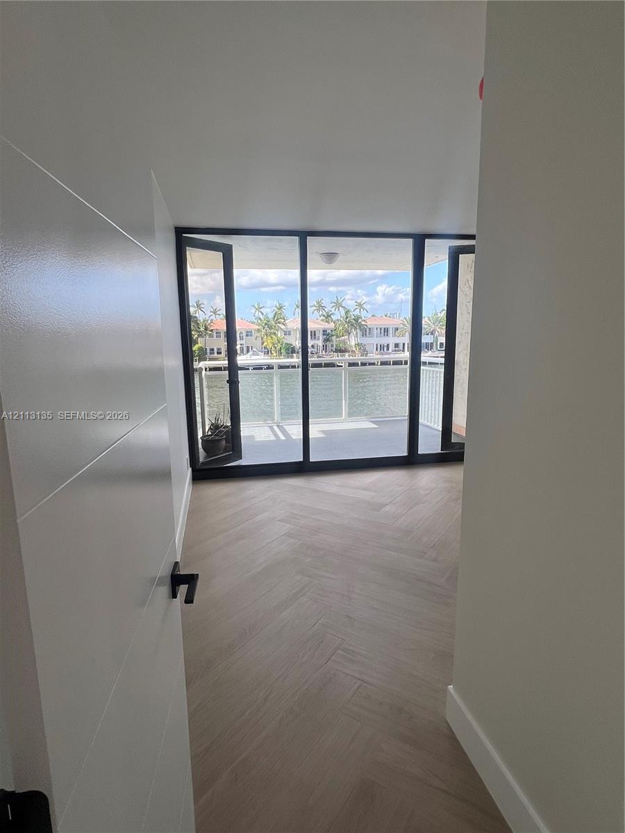 Herringbone-patterned wood-finish flooring, floor-to-ceiling glass panel doors, and a waterfront balcony with a railing