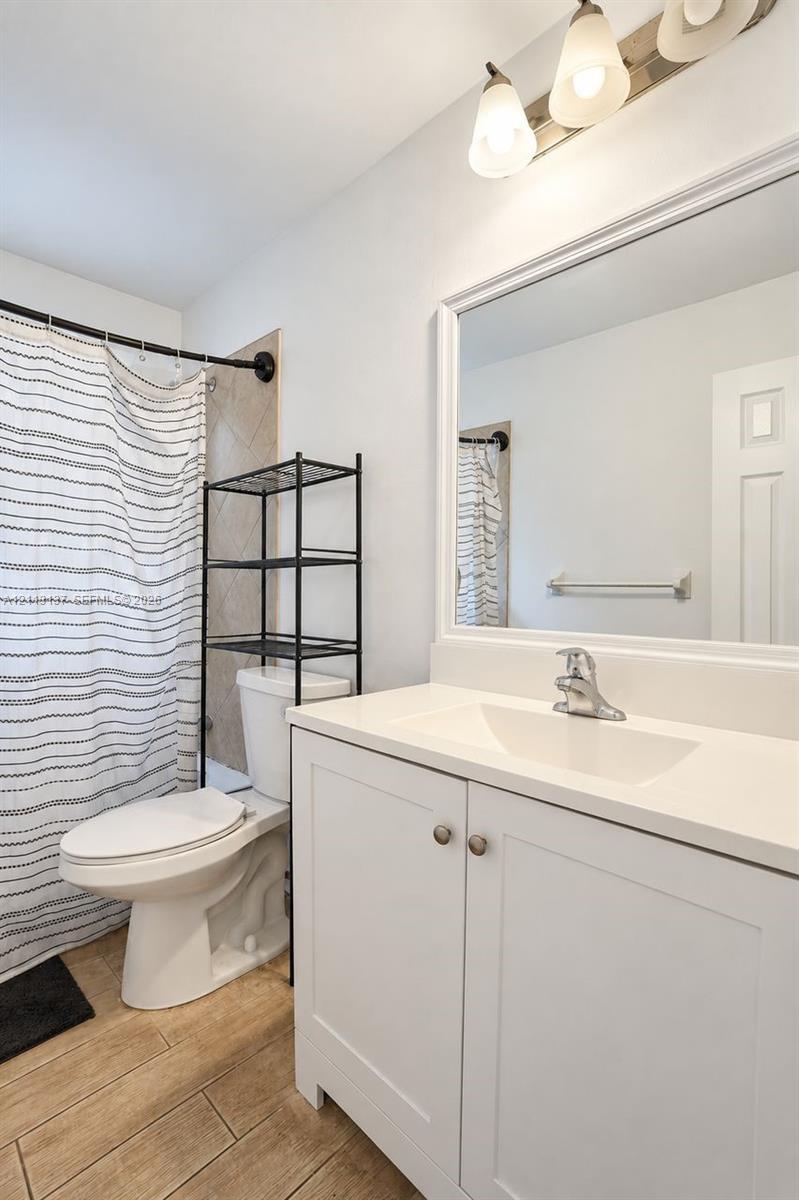 White vanity with a solid-surface countertop and integrated sink, framed wall mirror, three-light vanity fixture, and wood-finish flooring White vanity with a solid-surface countertop and integrated sink, framed wall mirror, three-light vanity fixture, and wood-finish flooring