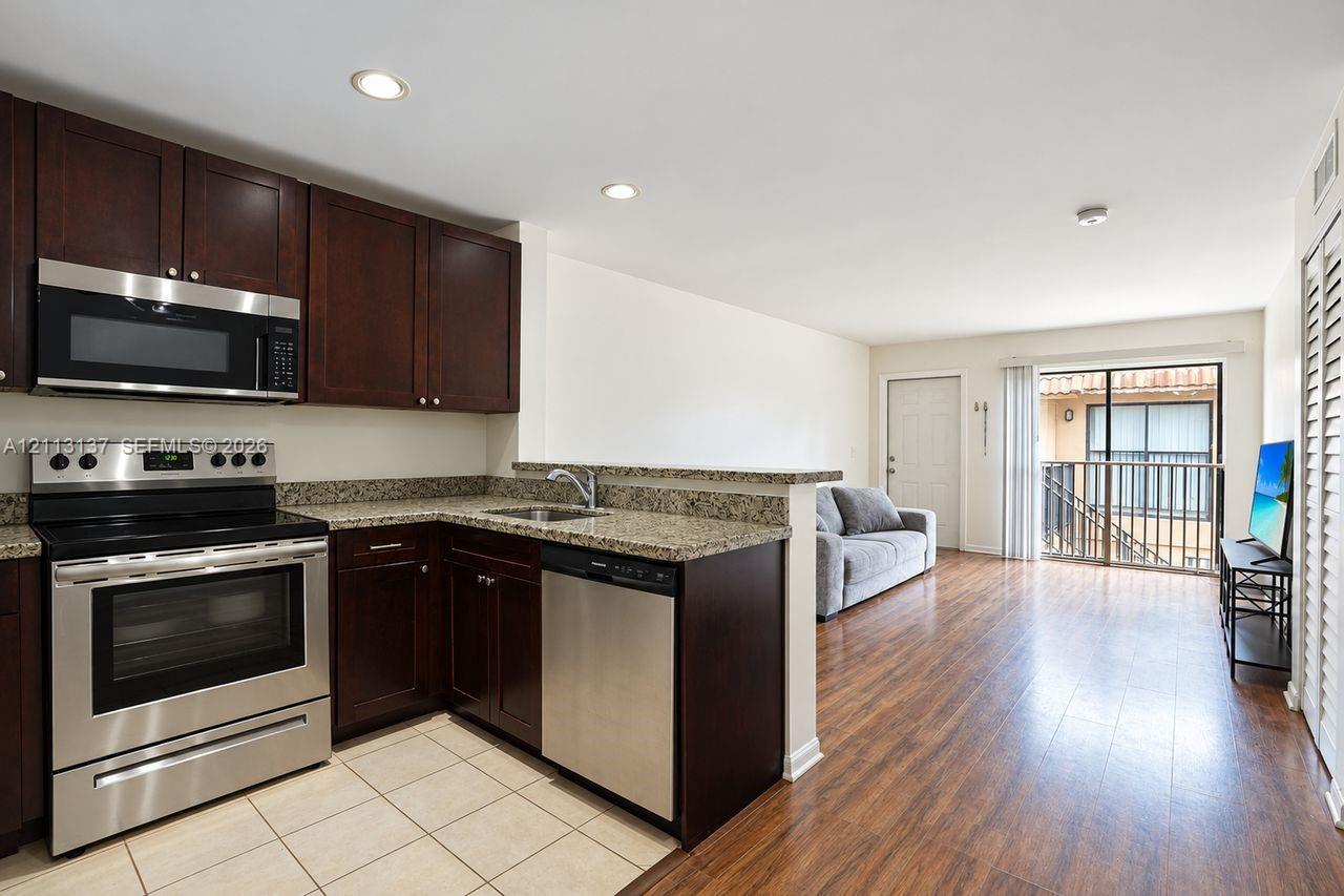 Kitchen featuring dark wood cabinetry, stone-finish countertops, stainless steel appliances, and tile flooring, paired with wood-finish flooring and a private balcony Kitchen featuring dark wood cabinetry, stone-finish countertops, stainless steel appliances, and tile flooring, paired with wood-finish flooring and a private balcony