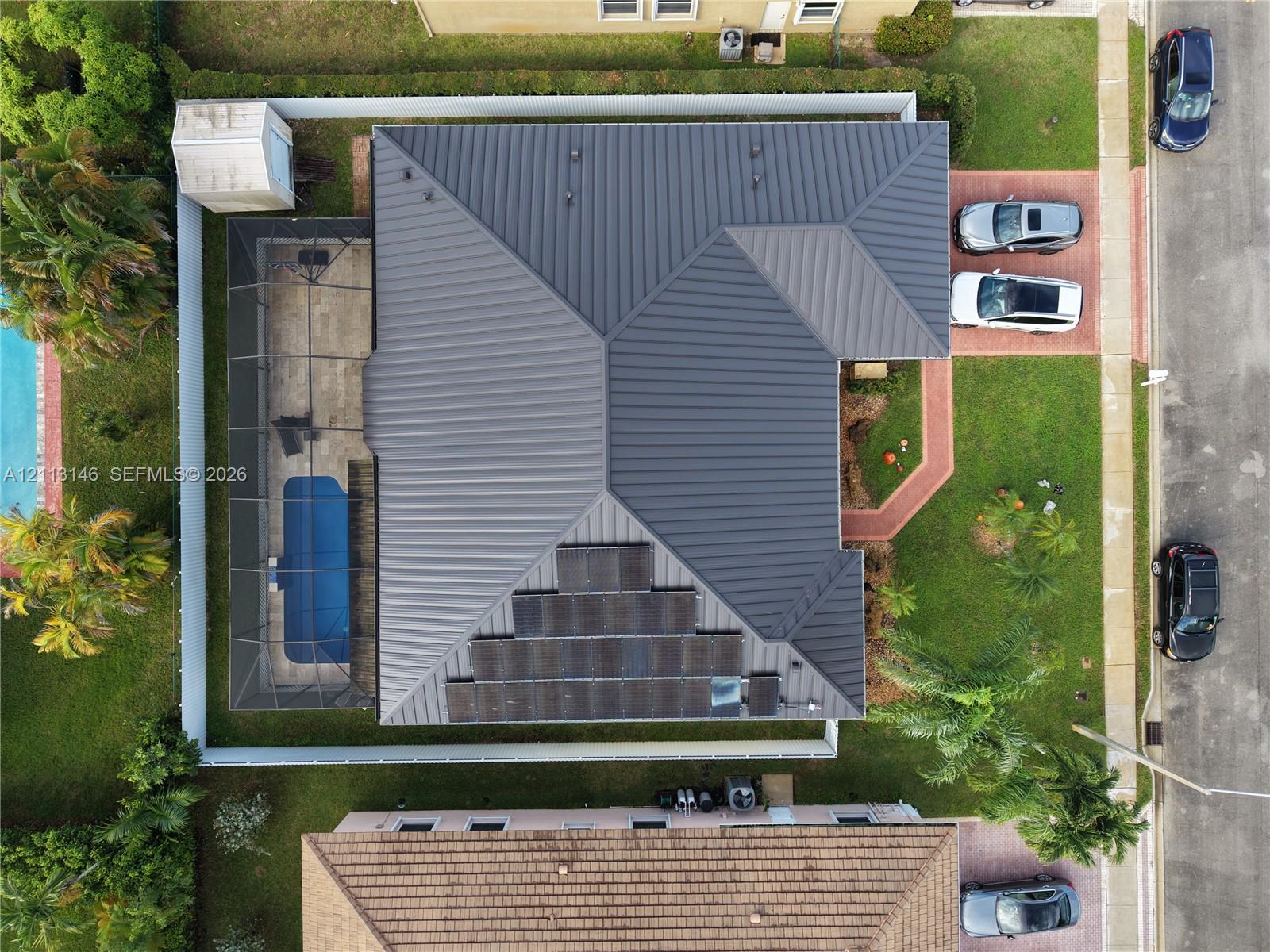Aerial perspective featuring a dark-toned metal roof, integrated solar panel array, screened-in swimming pool enclosure, paver driveway, and fenced perimeter Aerial perspective featuring a dark-toned metal roof, integrated solar panel array, screened-in swimming pool enclosure, paver driveway, and fenced perimeter