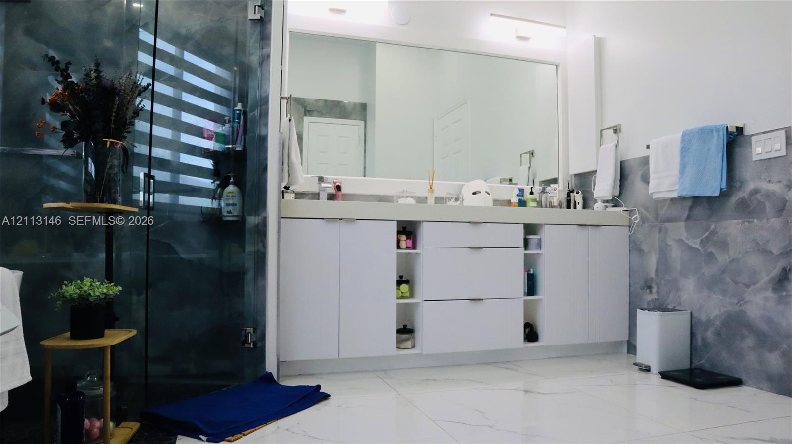 Dual-sink vanity with white cabinetry and open shelving, topped with a solid surface countertop, featuring a frameless glass shower enclosure and oversized tile flooring