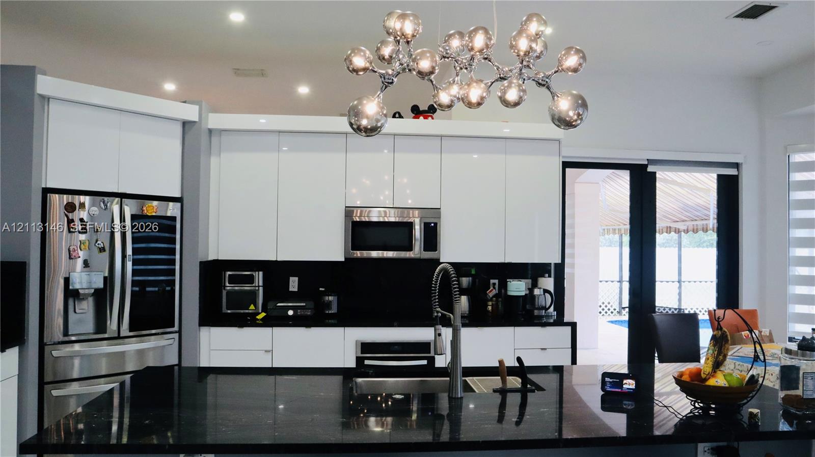 Kitchen featuring high-gloss white cabinetry, black stone countertops, built-in stainless steel appliances, a center island with a chrome gooseneck faucet, and a contemporary multi-light chrome chandelier Kitchen featuring high-gloss white cabinetry, black stone countertops, built-in stainless steel appliances, a center island with a chrome gooseneck faucet, and a contemporary multi-light chrome chandelier