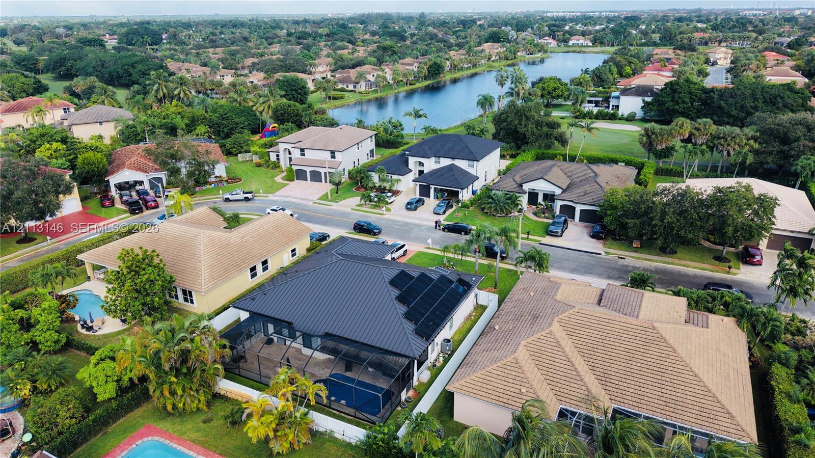 Aerial view of a residential property featuring a metal roof, integrated solar panels, a screened-in rear patio, and a private swimming pool, situated in a community with water features and established vegetation Aerial view of a residential property featuring a metal roof, integrated solar panels, a screened-in rear patio, and a private swimming pool, situated in a community with water features and established vegetation
