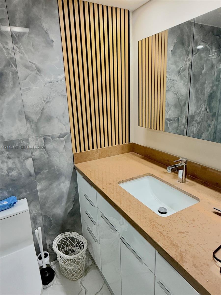 Custom vanity featuring a solid surface countertop with an integrated basin, high-gloss white cabinetry, vertical wood-slat accent wall, and large-format gray patterned wall tile