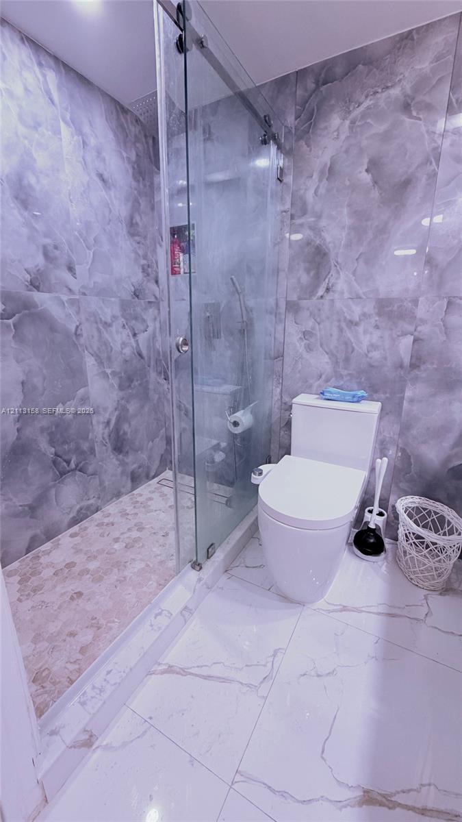 Walk-in shower with a sliding glass door, floor-to-ceiling gray-toned wall tiling, pebble-finish shower flooring, and oversized white floor tile with gray veining