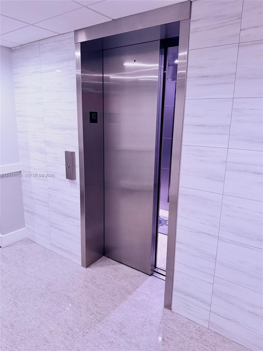 Stainless steel elevator with a brushed metal frame, surrounded by large-format, horizontal-vein tile wall cladding and a polished stone-finish floor Stainless steel elevator with a brushed metal frame, surrounded by large-format, horizontal-vein tile wall cladding and a polished stone-finish floor