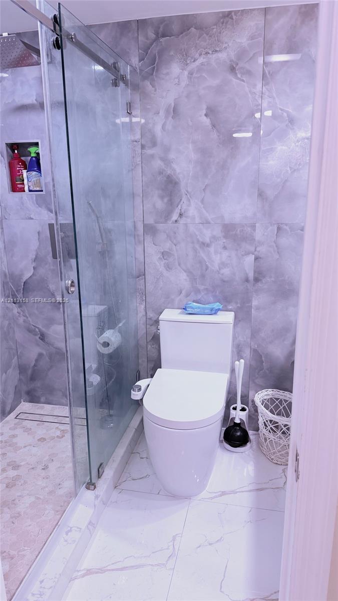 Walk-in shower with a sliding glass door, linear drain, built-in wall niche, and large-format gray stone-finish wall tile, complemented by white large-format stone-finish floor tile and a dual-flush toilet